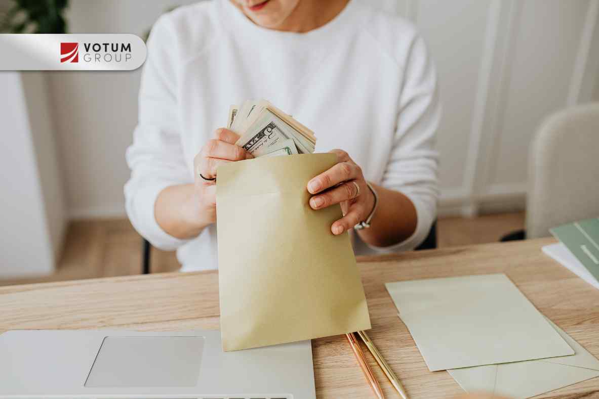 Zbliżenie na osobę w białym swetrze, która wkłada plik banknotów, do brązowej koperty na drewnianym biurku z otwartym laptopem
