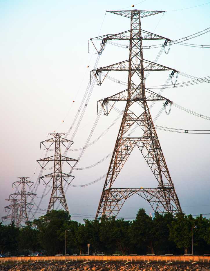 linie-energetyczne-odszkodowanie-pomoc Duży, masywny słup linii wysokiego napięcia wraz z kilkoma mniejszymi w tle, rozciągający się nad rzędem drzew.