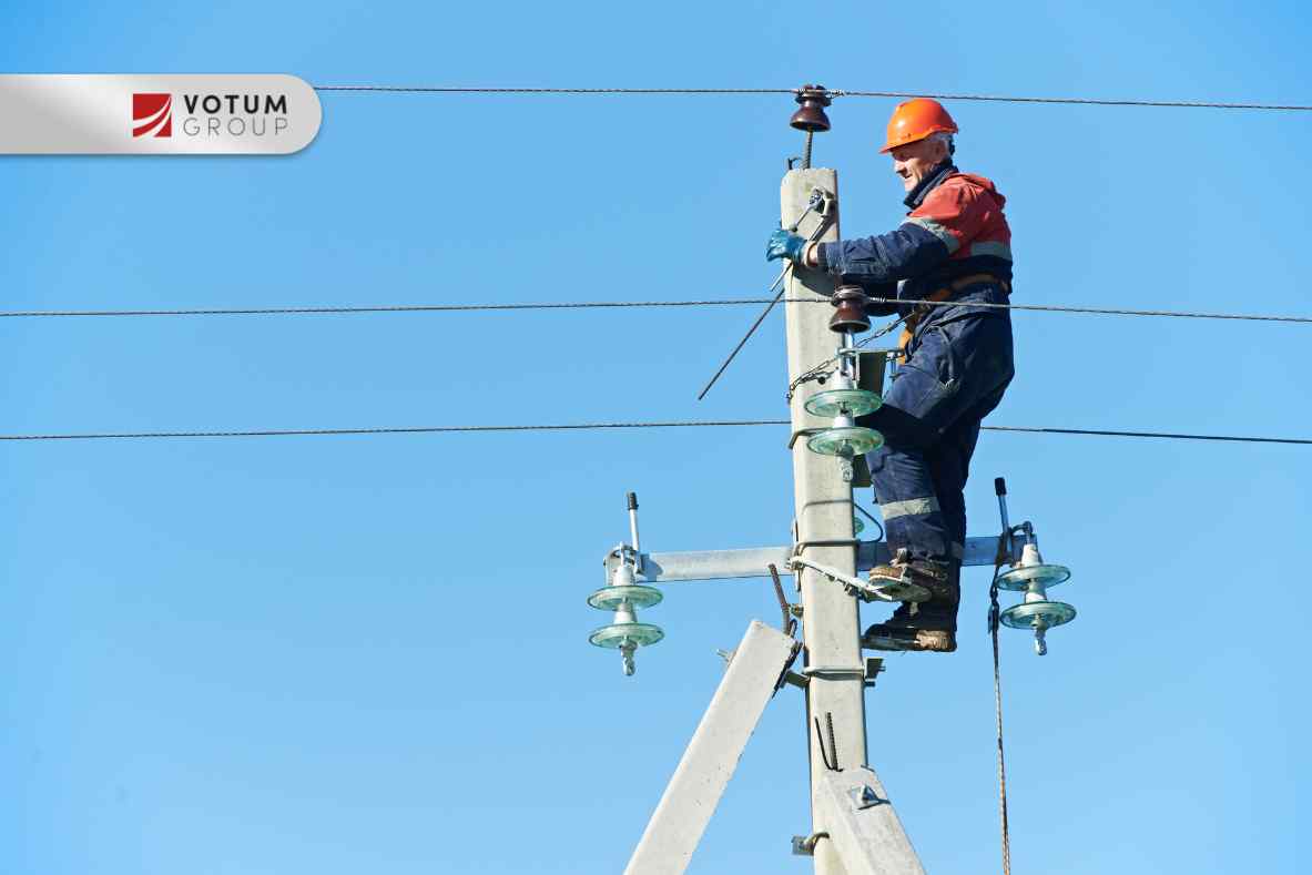 Mężczyzna w pomarańczowym kasku i kombinezonie wisi na betonowym słupie energetycznym, pracując przy izolatorach i liniach, na tle jasnego, błękitnego nieba.