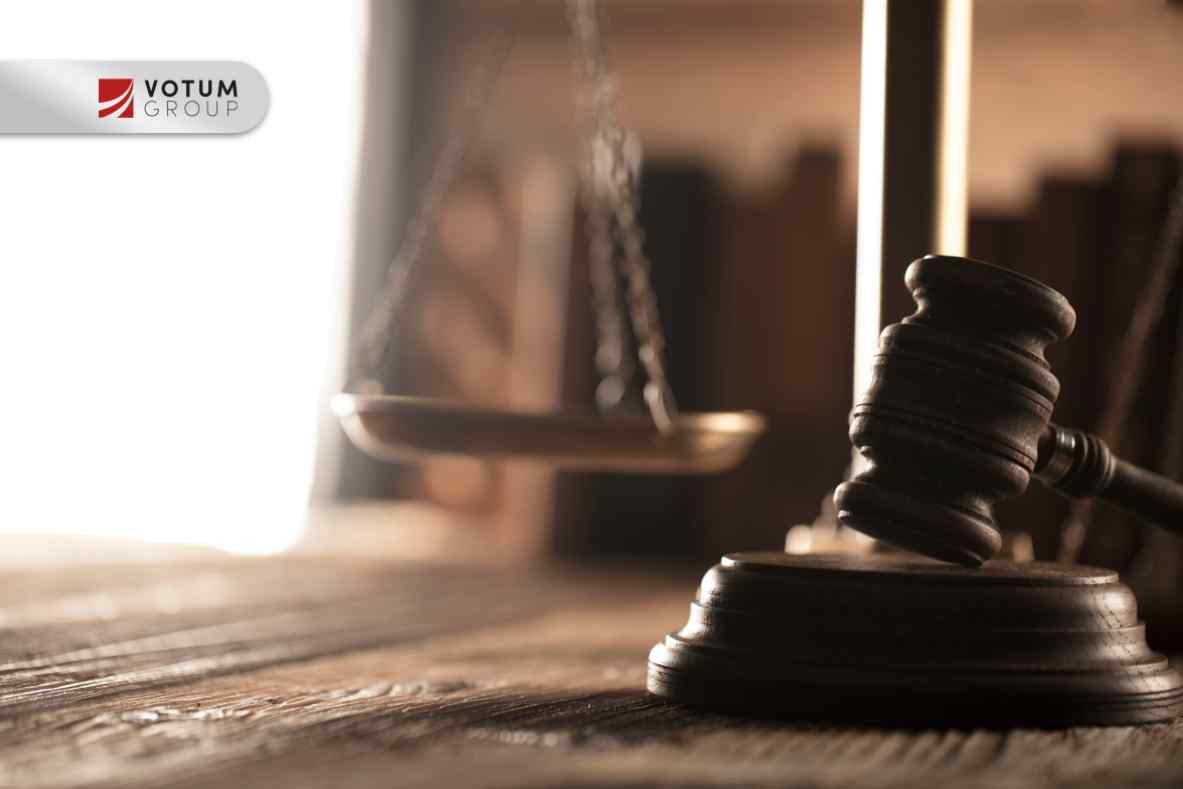 Drewniana powierzchnia stołu, na której leży ciemny młotek sędziowski (gavel) oraz mosiężna szala sprawiedliwości w tle.