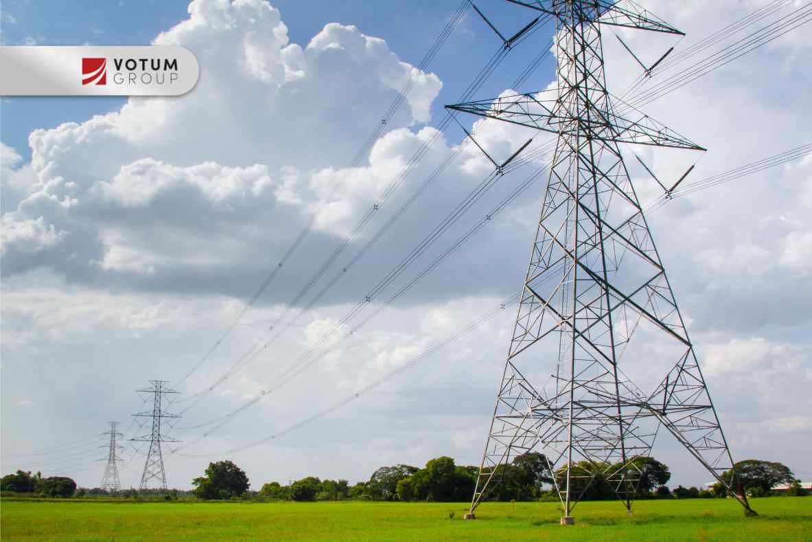 Widok na liczne, wysokie metalowe słupy wysokiego napięcia na zielonej łące, na tle błękitnego nieba z dużymi białymi chmurami