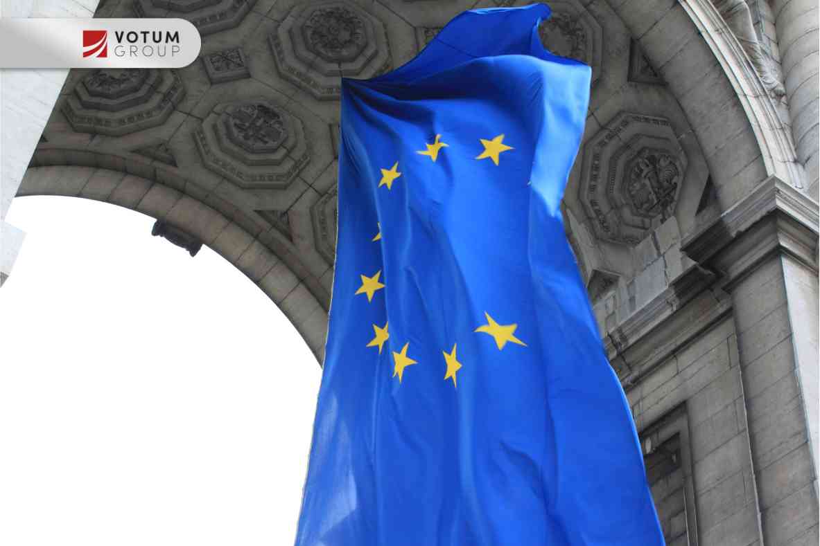 Pionowa, niebieska flaga Unii Europejskiej z żółtymi gwiazdami, powiewająca na tle zabytkowego, kamiennego łuku o bogatych zdobieniach architektonicznych.