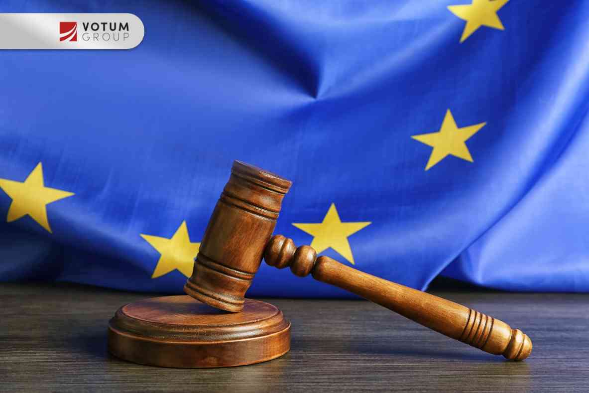 Drewniany młotek sędziowski spoczywający na podstawce na ciemnym blacie, w tle powiewająca flaga Unii Europejskiej z żółtymi gwiazdami.