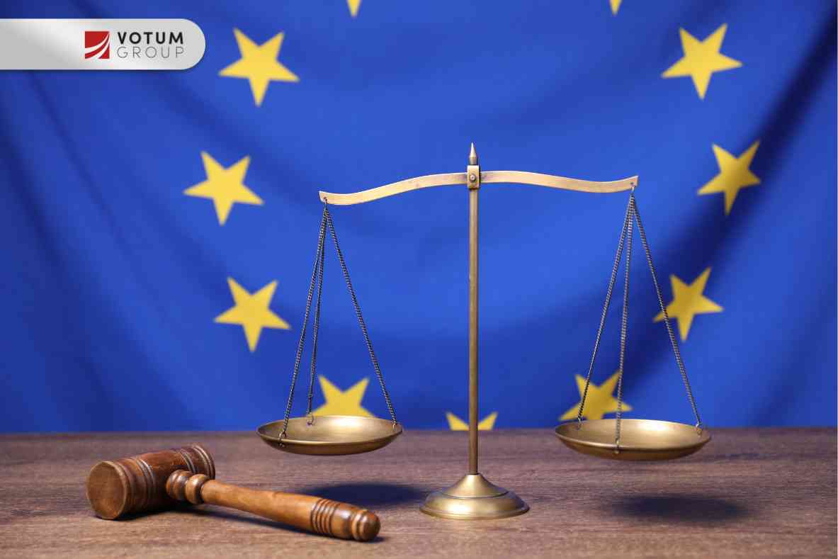 Złota waga szali stoi w centrum na drewnianym stole, obok niej leży drewniany młotek sędziowski. Całość na tle niebieskiej flagi Unii Europejskiej z żółtymi gwiazdami.