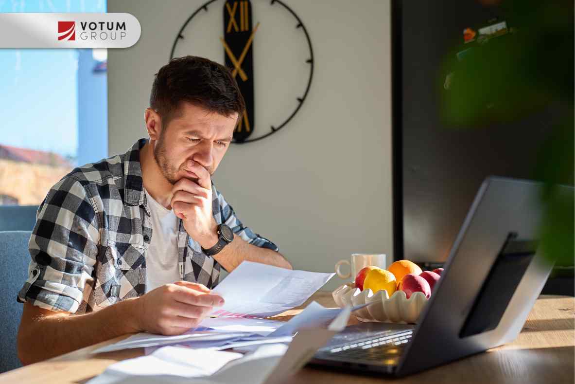 Mężczyzna w koszuli w kratę siedzi przy biurku z laptopem, podpierając brodę w geście zamyślenia; analizuje rozłożone przed nim dokumenty bankowe.