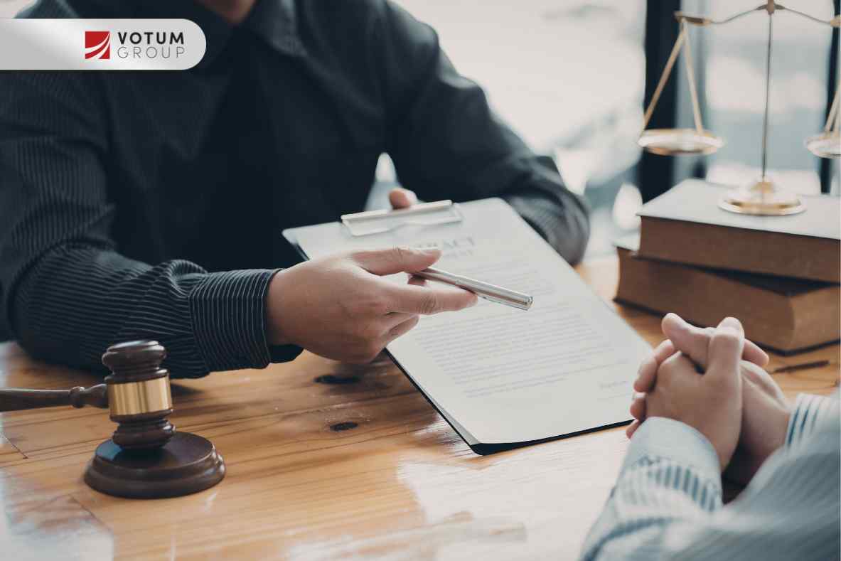 Prawnik przekazuje klientowi dokument do podpisu na drewnianym biurku, na którym stoją również młotek sędziowski, waga sprawiedliwości oraz stos książek.