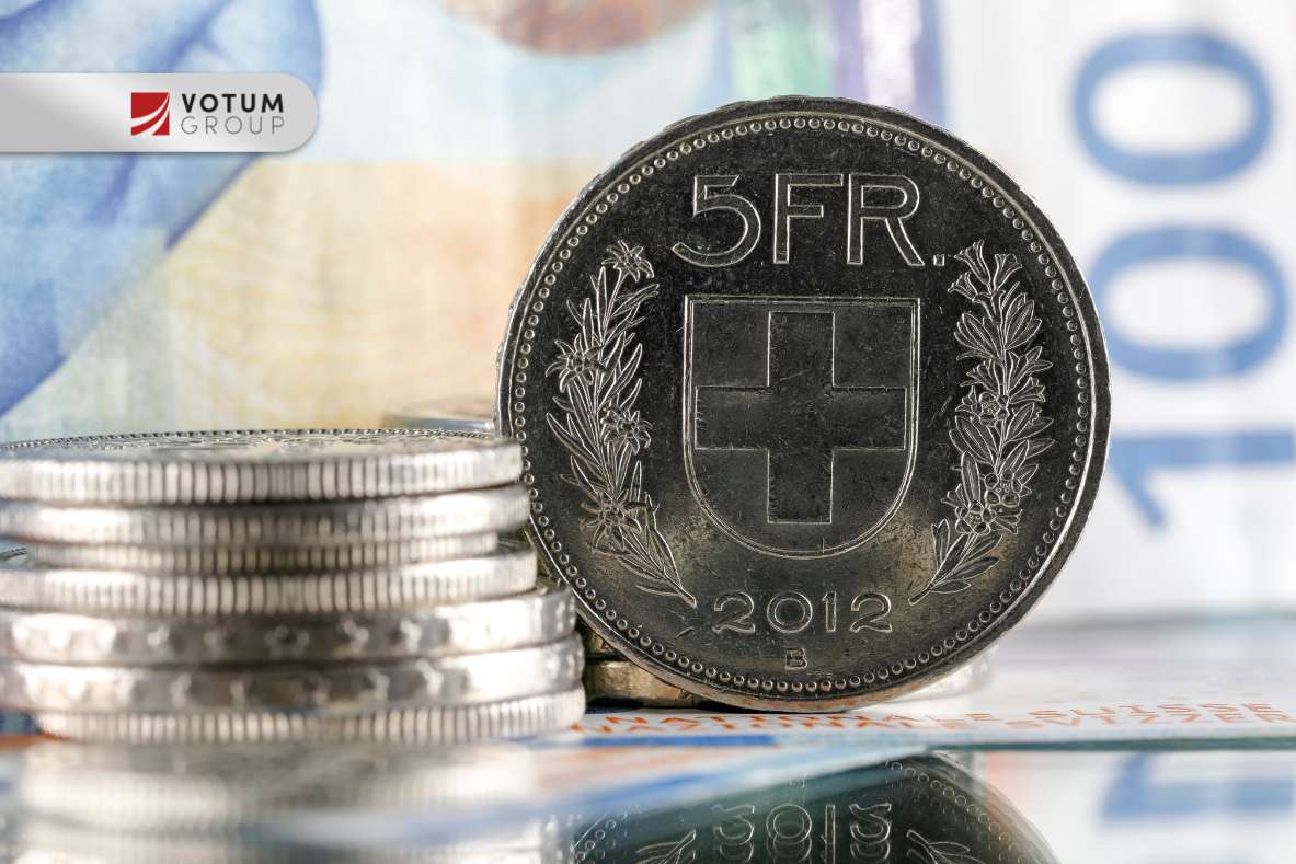 Close-up of a 5 FR coin (2012) standing beside a stack of coins on a reflective surface; Votum Group logo visible in the corner.