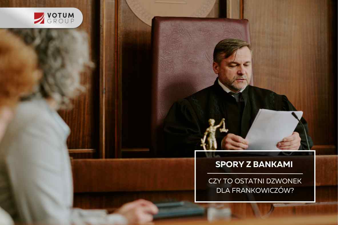 Sędzia w czarnej todze siedzi przy stole sędziowskim w sali rozpraw, czytając dokumenty; na pierwszym planie widać rozmyte sylwetki osób uczestniczących w rozprawie.