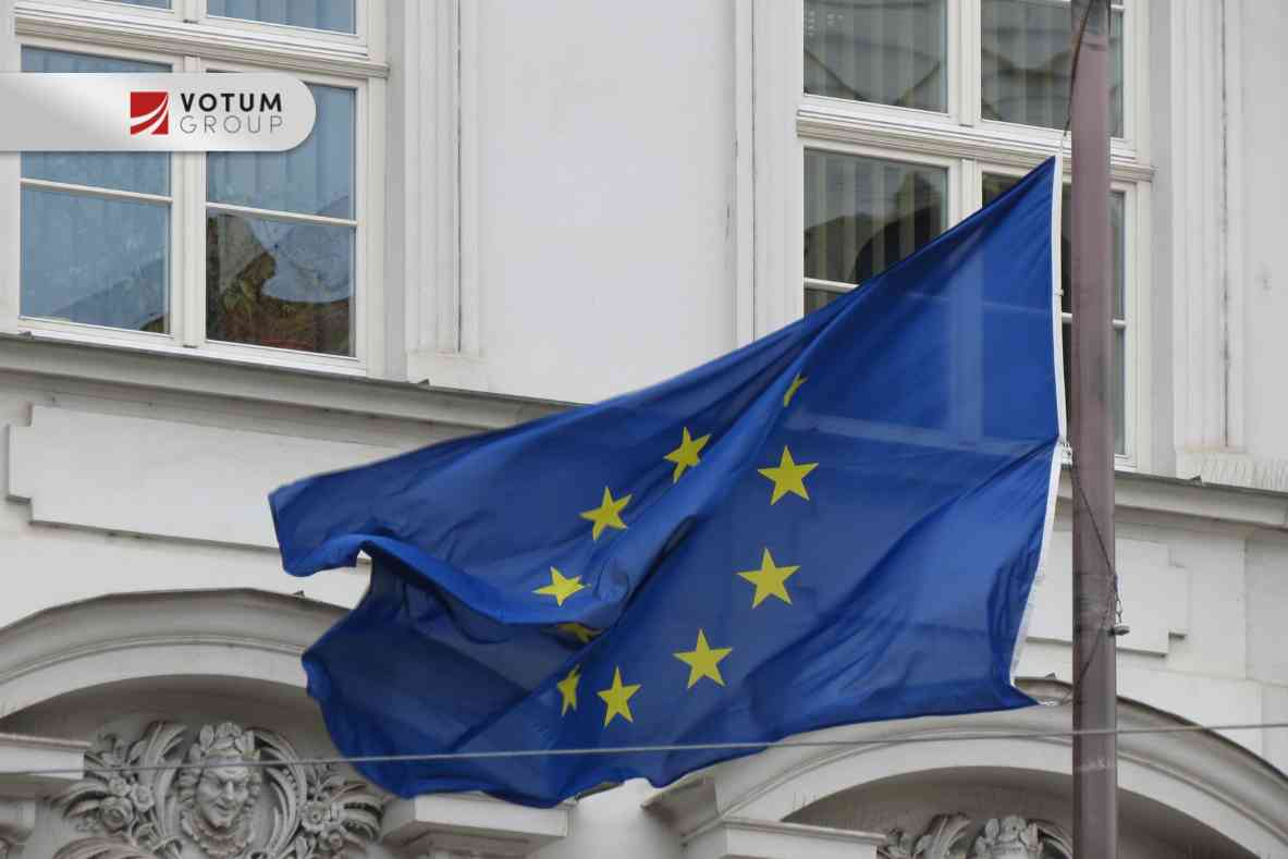 European Union flag waving in front of a white building with a Votum Group sign visible above the windows.