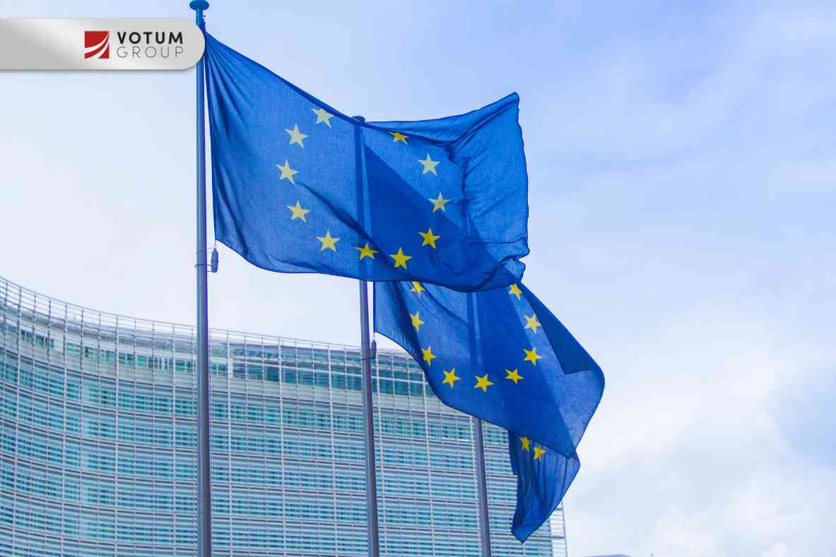 Złota waga szali stoi w centrum na drewnianym stole, obok niej leży drewniany młotek sędziowski. Całość na tle niebieskiej flagi Unii Europejskiej z żółtymi gwiazdami.