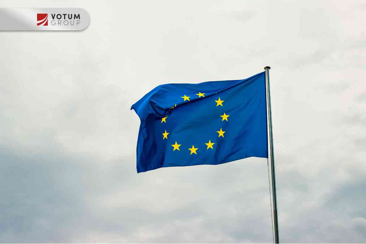 Drewniany młotek sędziowski spoczywający na podstawce na ciemnym blacie, w tle powiewająca flaga Unii Europejskiej z żółtymi gwiazdami.