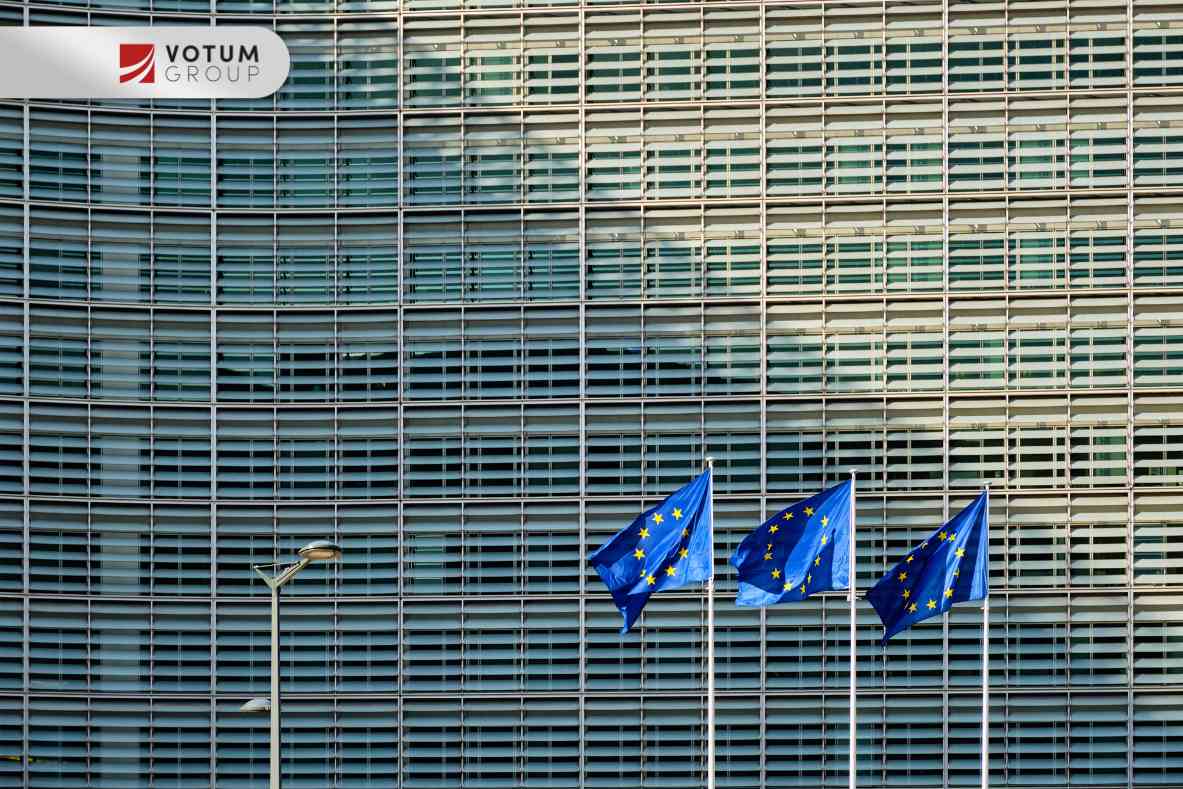 Trzy flagi Unii Europejskiej powiewające na masztach przed nowoczesnym biurowcem o szklanej, pasiastej elewacji.