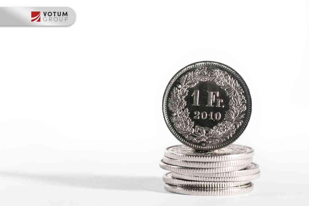 Close-up of a 5 FR coin (2012) standing beside a stack of coins on a reflective surface; Votum Group logo visible in the corner.