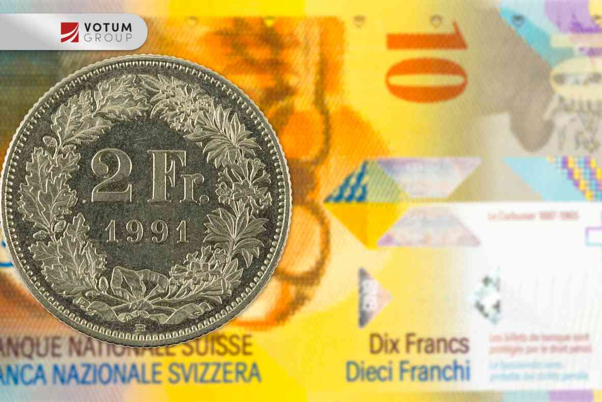 Close-up of a 5 FR coin (2012) standing beside a stack of coins on a reflective surface; Votum Group logo visible in the corner.