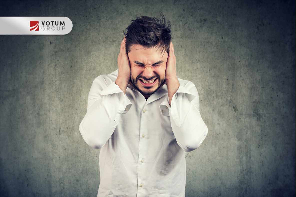 Man in a white shirt covers his ears and grimaces in distress against a gray textured background, with a VOTUM GROUP logo in the top-left corner.